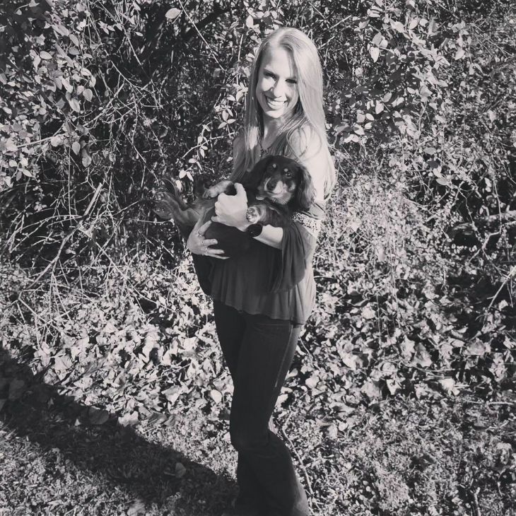 Smiling woman standing outdoors on a leafy path, holding a relaxed long-haired Dachshund in her arms